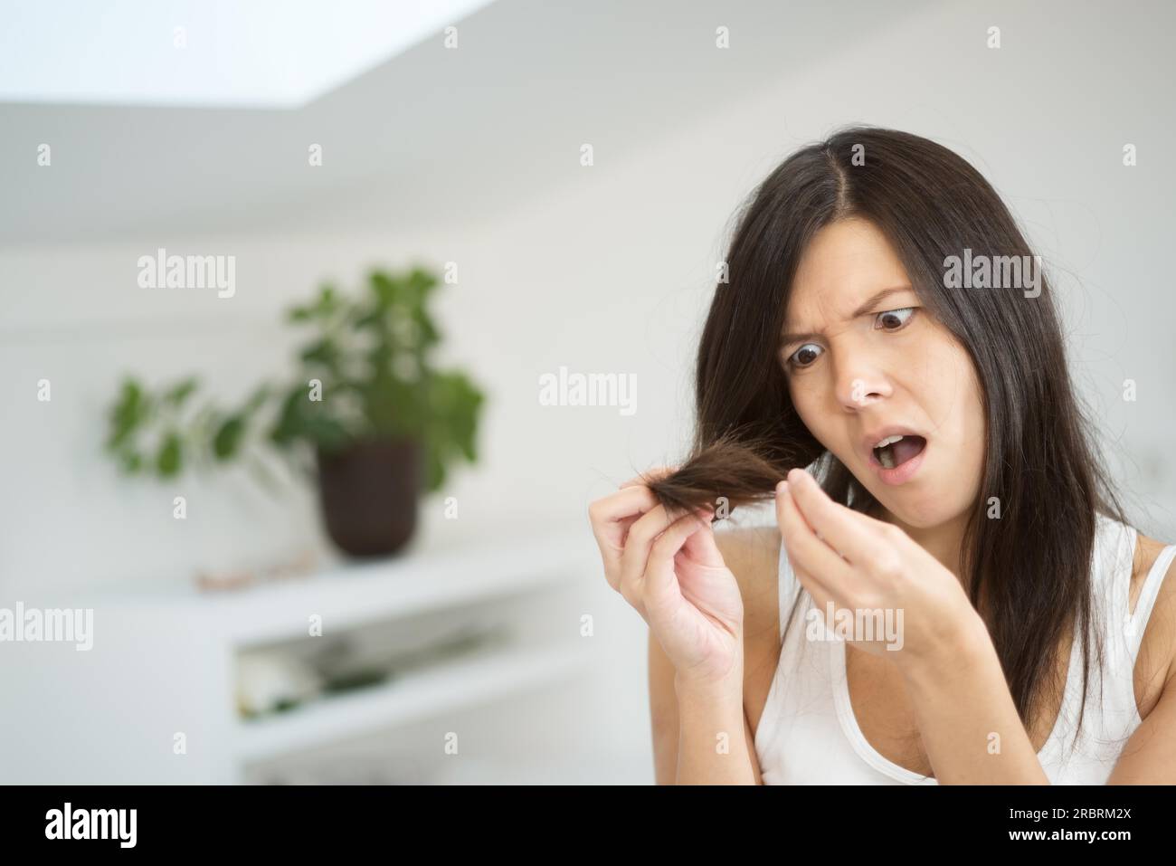 Eine Frau, die die Enden ihrer langen brünetten Haare auf ihren Zustand überprüft und die Enden mit einem Grimace im Gesicht, während sie im Badezimmer steht Stockfoto