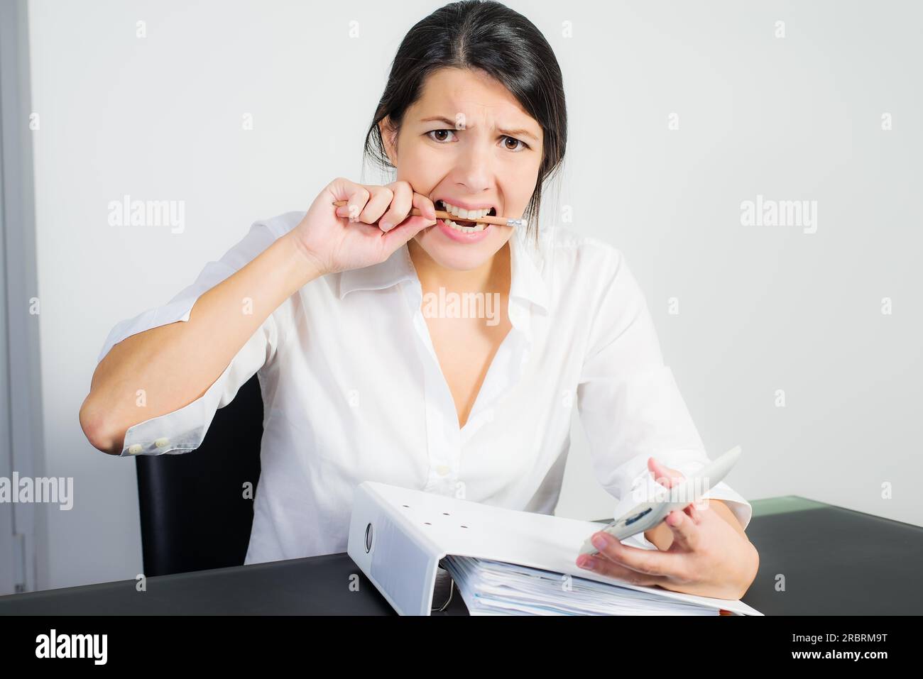 Geschäftsfrau mit einem verzweifelten Ausdruck beißen ihre Feder in Frustration, Angst oder Wut, als sie an ihrem Schreibtisch mit einem Taschenrechner in der Hand sitzt Stockfoto
