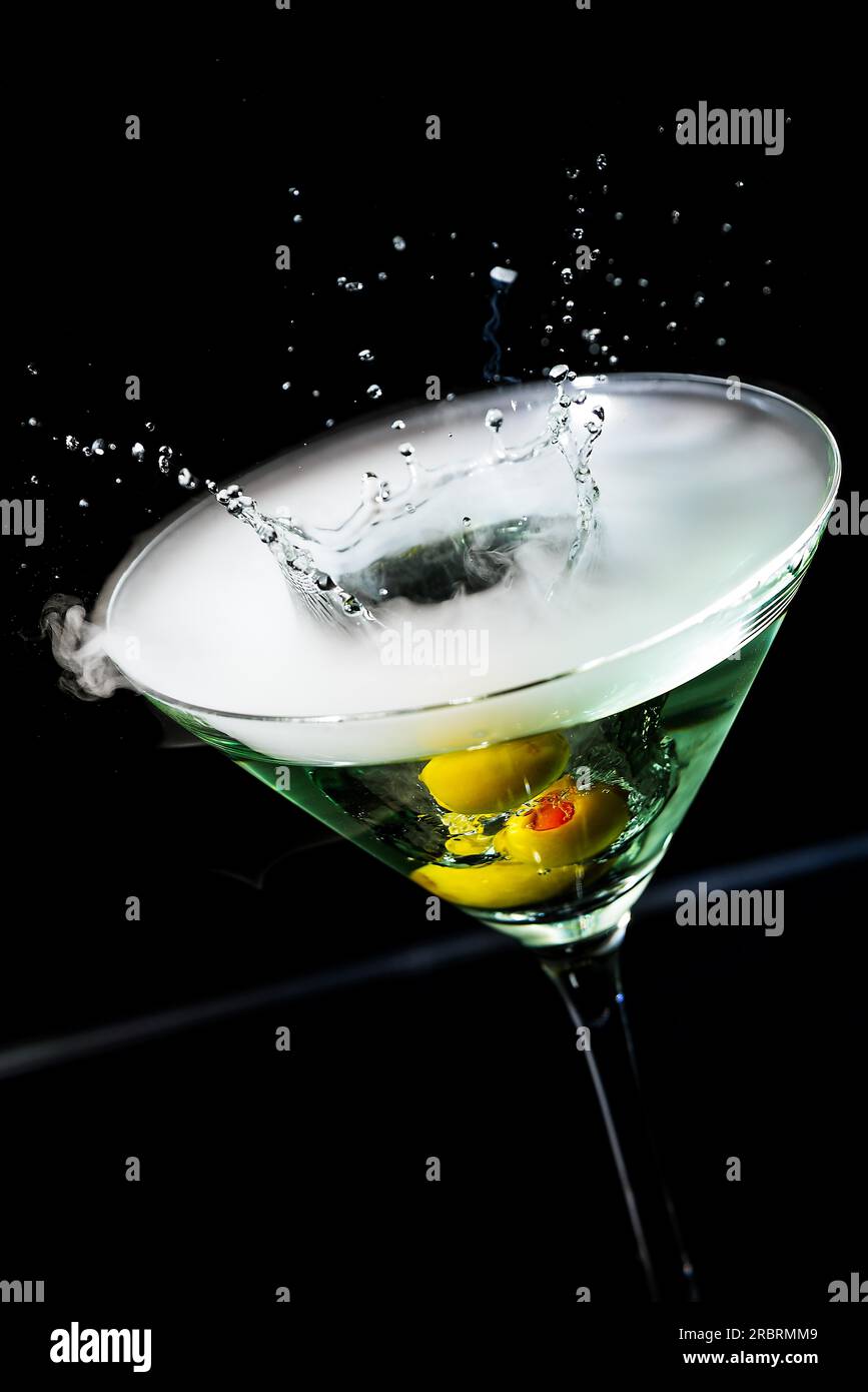 Nahaufnahme der konischen Schüssel eines eleganten Cocktailglases mit Oliven, die in einen martini-Cocktail mit suspendiertem Spray und Tröpfchen über einem spritzen Stockfoto