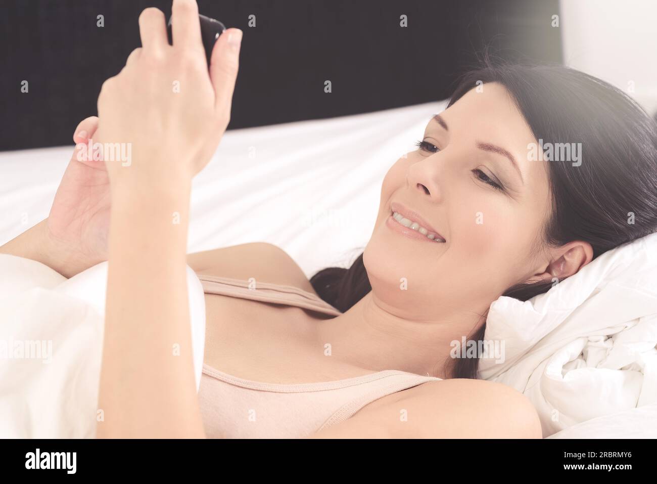 Attraktive junge Frau liegt im Bett und las auf Sms auf ihrem Mobiltelefon lächelnd mit Freude über die Nachricht, Closeup-Seitenansicht Stockfoto
