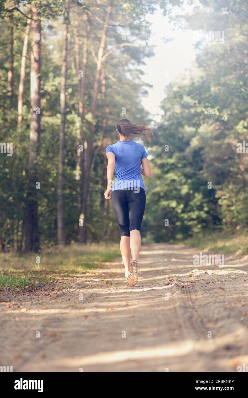 Sportliche junge Frau in Sportbekleidung, die in einem Fitness- und Wellnesskonzept von der Kamera weg auf einer Waldstrecke joggt Stockfoto