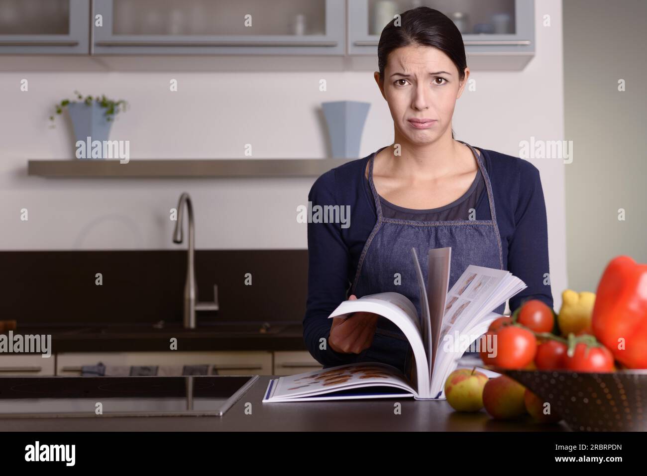 Traurige junge Frau trägt Schürze und hält ein Rezeptbuch in der Küche, während sie in die Kamera schaut Stockfoto