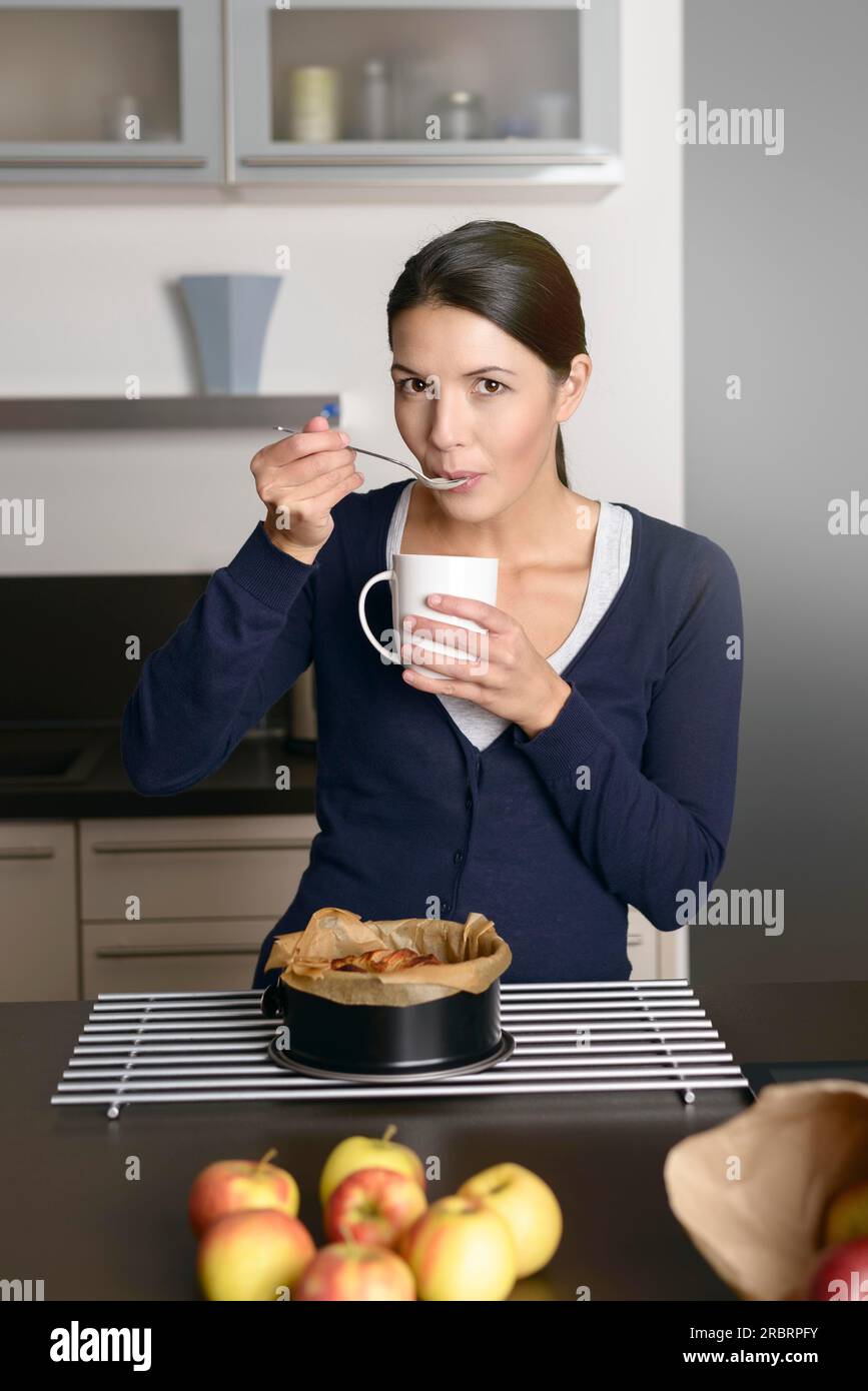 Lächelnde junge Frau, die Apfelkuchen backt, steht mit einer frisch aus dem Ofen entfernten Backform auf einem Kühlregal, während sie mit einem Löffel probiert Stockfoto