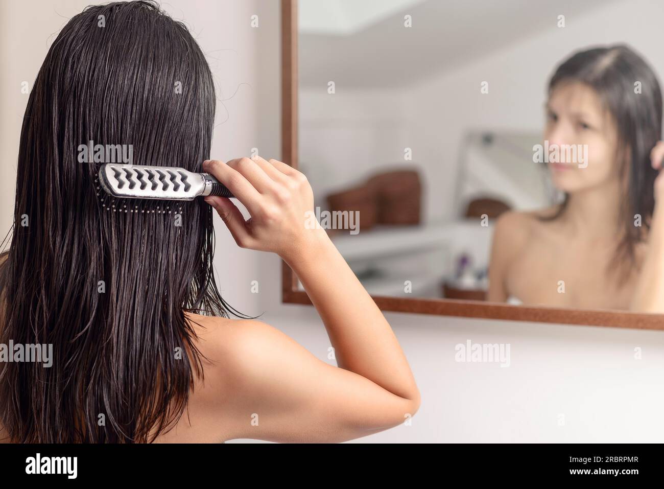 Junge Frau, die im Spiegel steht, während sie ihr nasses Haar nach dem Waschen mit einer Haarbürste bürstet, Blick von hinten Stockfoto