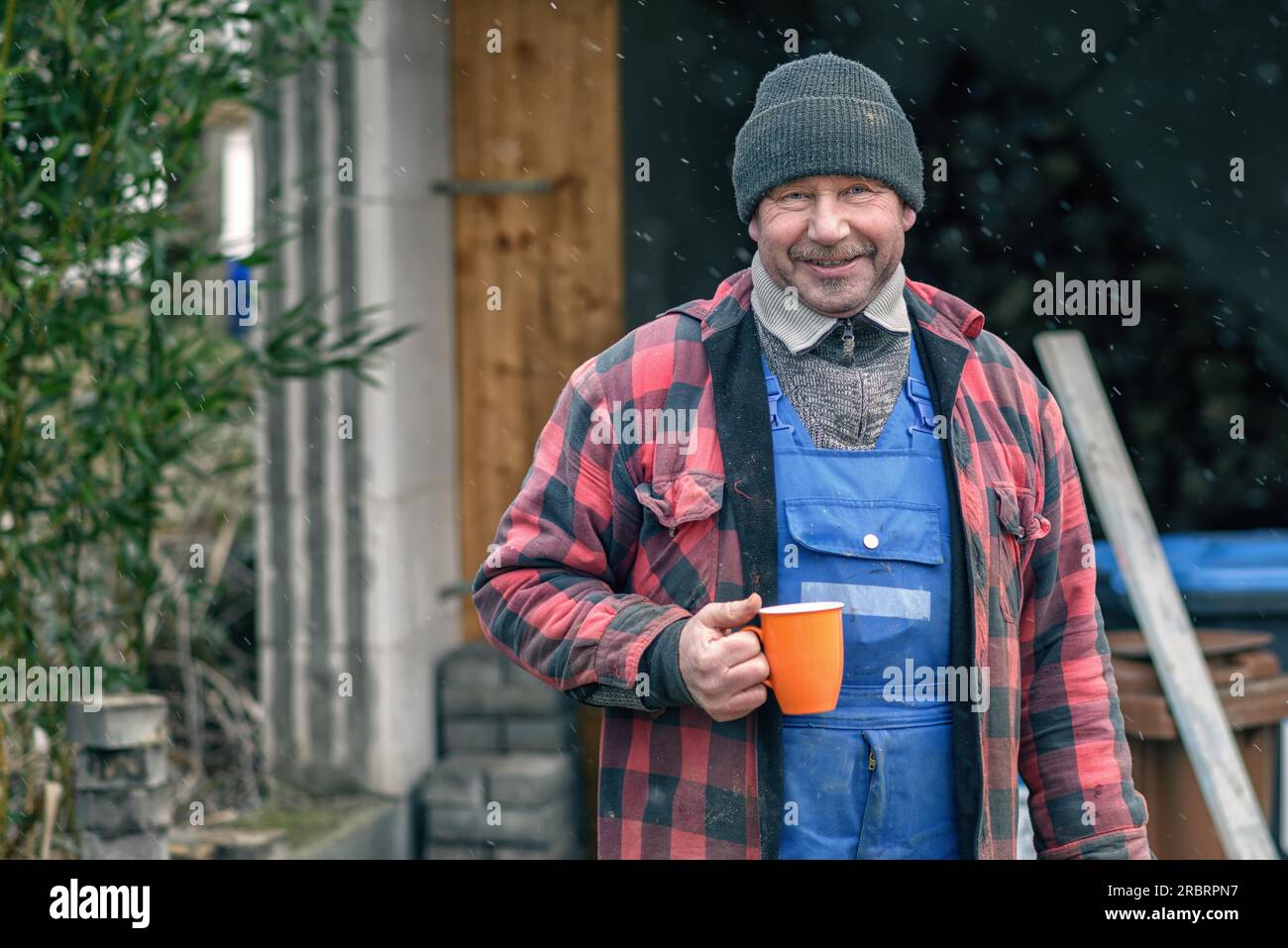 Glücklicher Mann in einem gestrickte Wolle Beanie, Jersey und Jacke stand draußen in den schneereichen Winter Wetter trinken heißen Kaffee aus einem hellen orange Becher Stockfoto