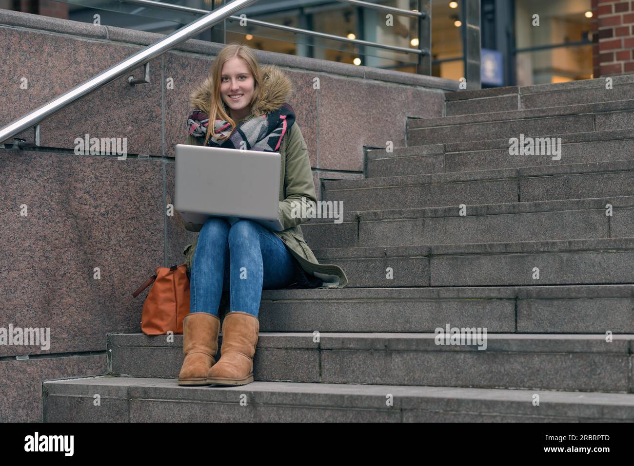 Hübsche junge Frau in einem eleganten, lässigen Winterkleid, die auf Treppen in der Stadt sitzt und ein Notebook auf den Knien hat, während sie auf das Internet zugreift oder sie lernt Stockfoto