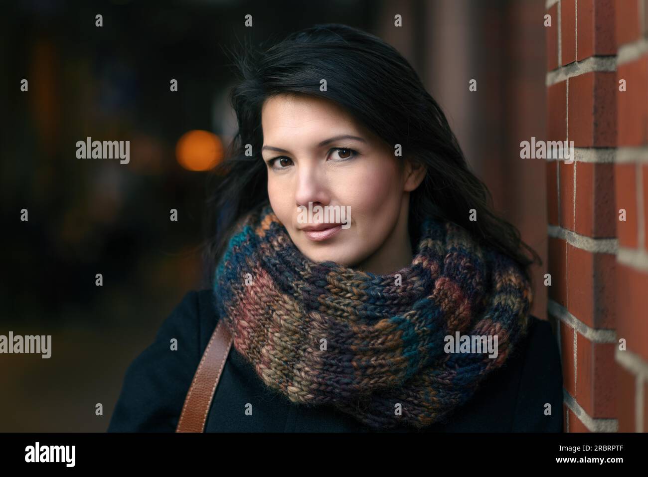 Stilvolle, hübsche junge Frau in Herbstmode, die sich auf eine rote Backsteinsäule lehnt und die Kamera anschaut Stockfoto