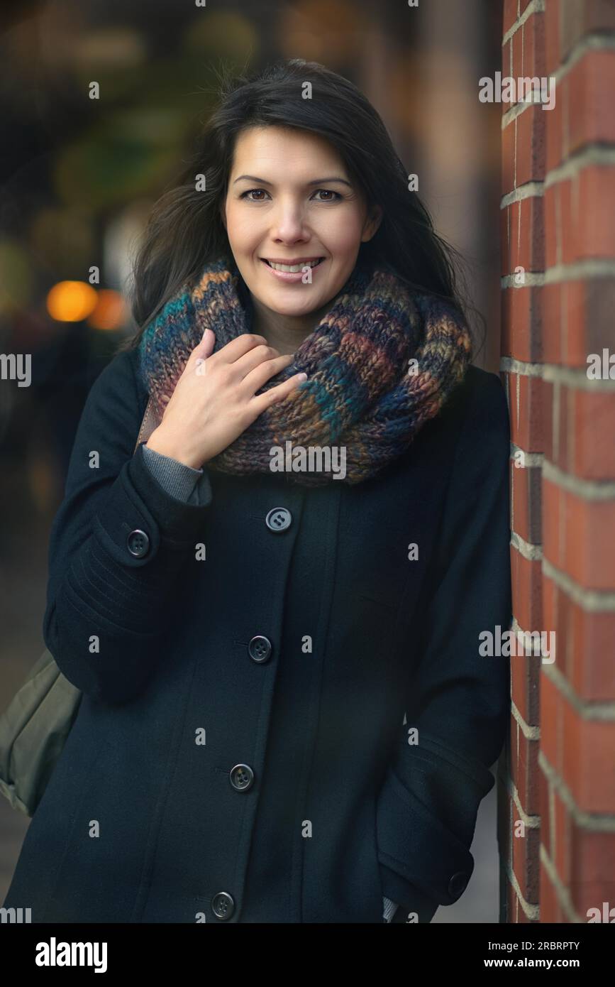 Stilvolle, hübsche junge Frau in Herbstmode, die sich auf eine rote Backsteinsäule lehnt und die Kamera anschaut Stockfoto