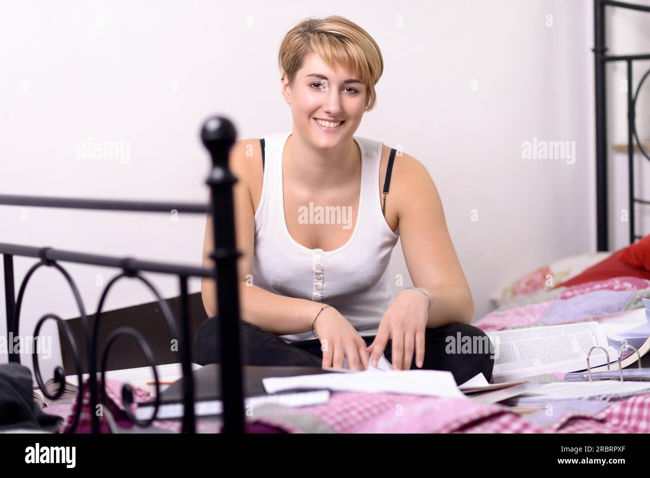 Sehen Sie sich eine lächelnde junge blonde Frau in lässiger Kleidung an, die mit gekreuzten Beinen auf ihrem Bett sitzt, während sie ein Buch scannt Stockfoto