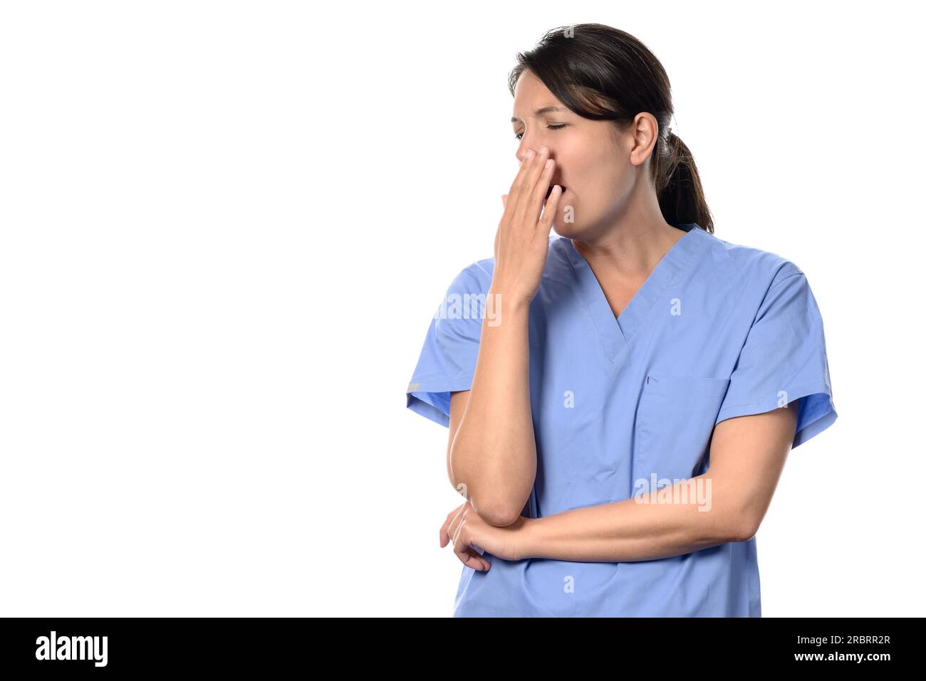 Erschöpfte, attraktive Ärztin oder Krankenschwester in blauen Kissen, die mit ihrer Hand an ihren Mund gähnte, einer langen, ermüdenden Schicht, isoliert auf weiß Stockfoto
