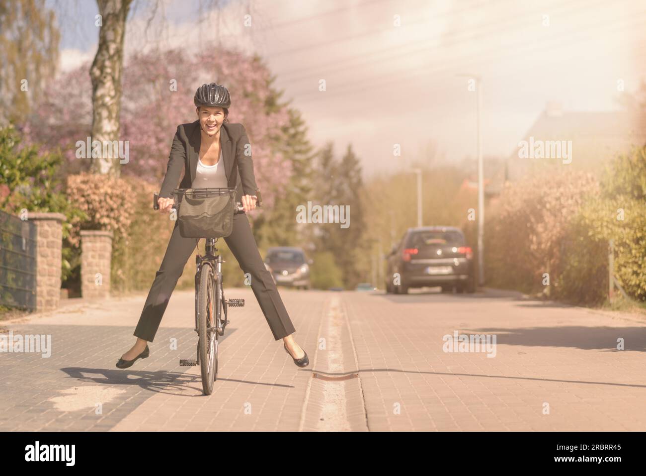 Eine stilvolle Frau in einem lockeren Anzug und einem Schutzhelm, die Spaß hat, auf ihrem Fahrrad zu arbeiten, ihre Füße in die Luft hebt und während der Küste balanciert Stockfoto