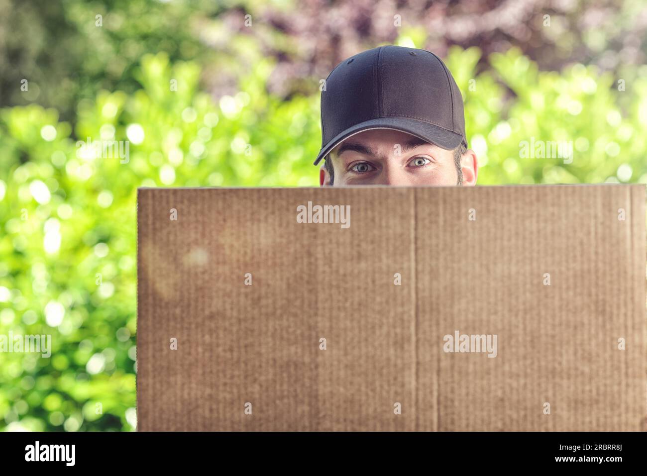 Ein Mann liefert einen großen Karton, der die untere Hälfte seines Gesichts bedeckt, sodass nur seine Augen und die Kappe vor einem frischen Hintergrund zu sehen sind Stockfoto