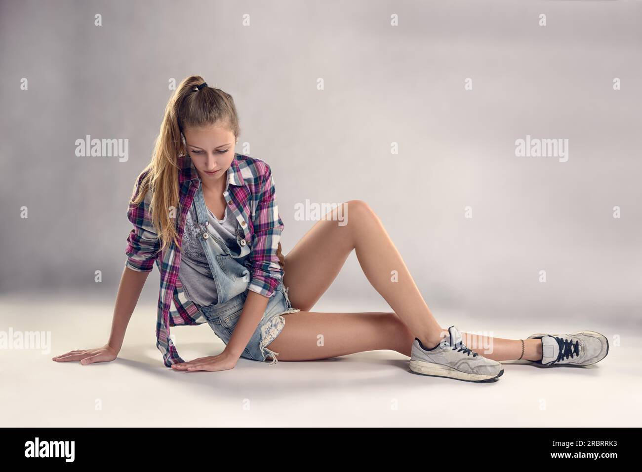 Stilvolle junge, moderne weibliche Tänzerin, die auf dem Boden sitzt, mit einem durchdachten Gesichtsausdruck vor grauem Wandhintergrund Stockfoto