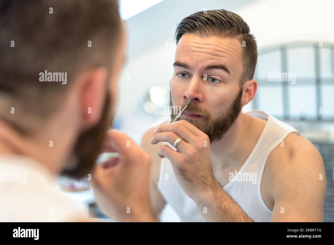 Gut aussehend bärtiger junger Mann stehend seine Nase Haare vor dem Spiegel im Badezimmer während seiner täglichen Fellpflege zupfen Stockfoto
