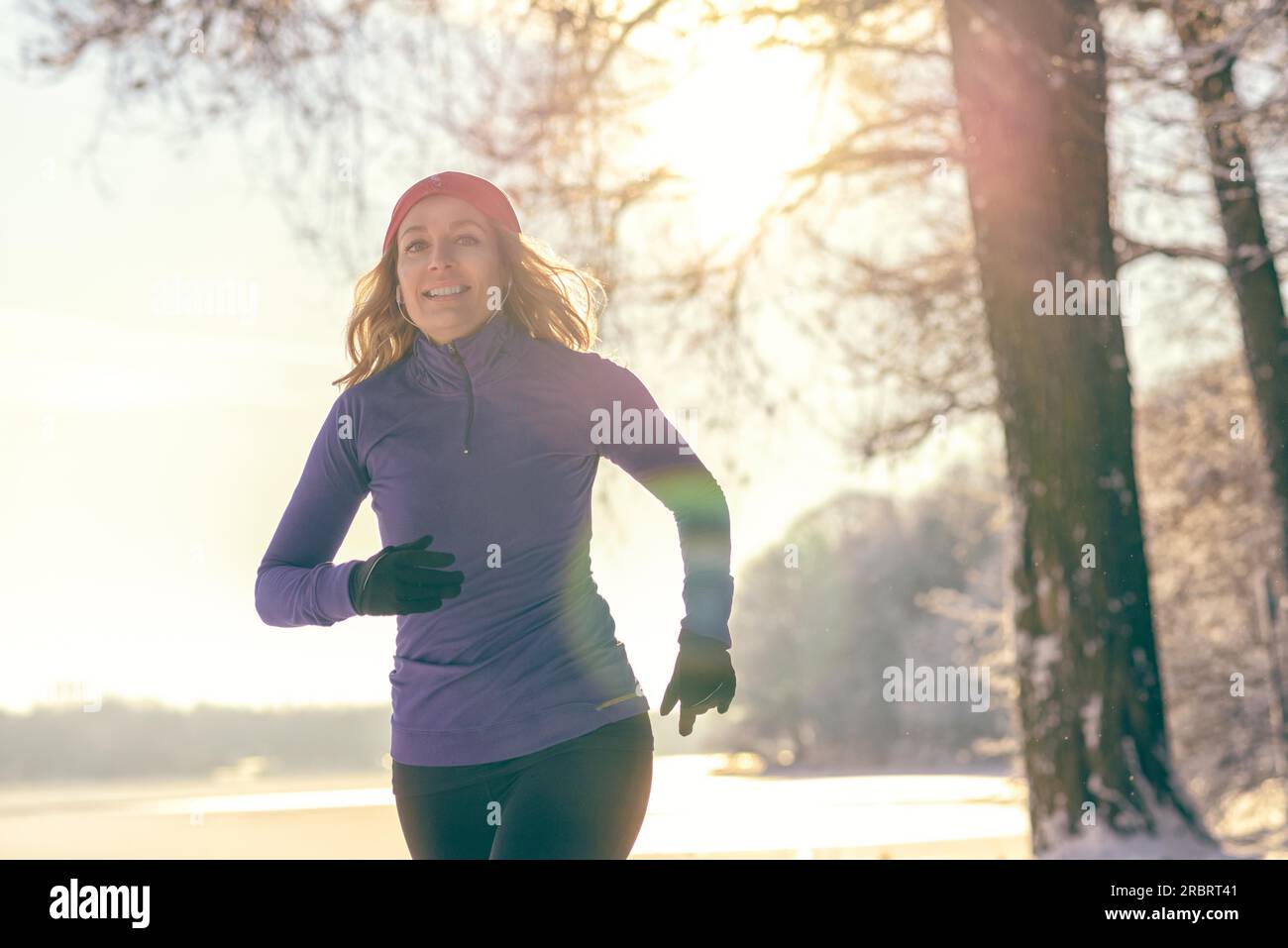 Ganzkörperaufnahme einer hübschen Athletikerin, die im Winter mit Kopfhörern und Winterkleidung joggt, die Kamera anlächelt, mit Kopierbereich Stockfoto