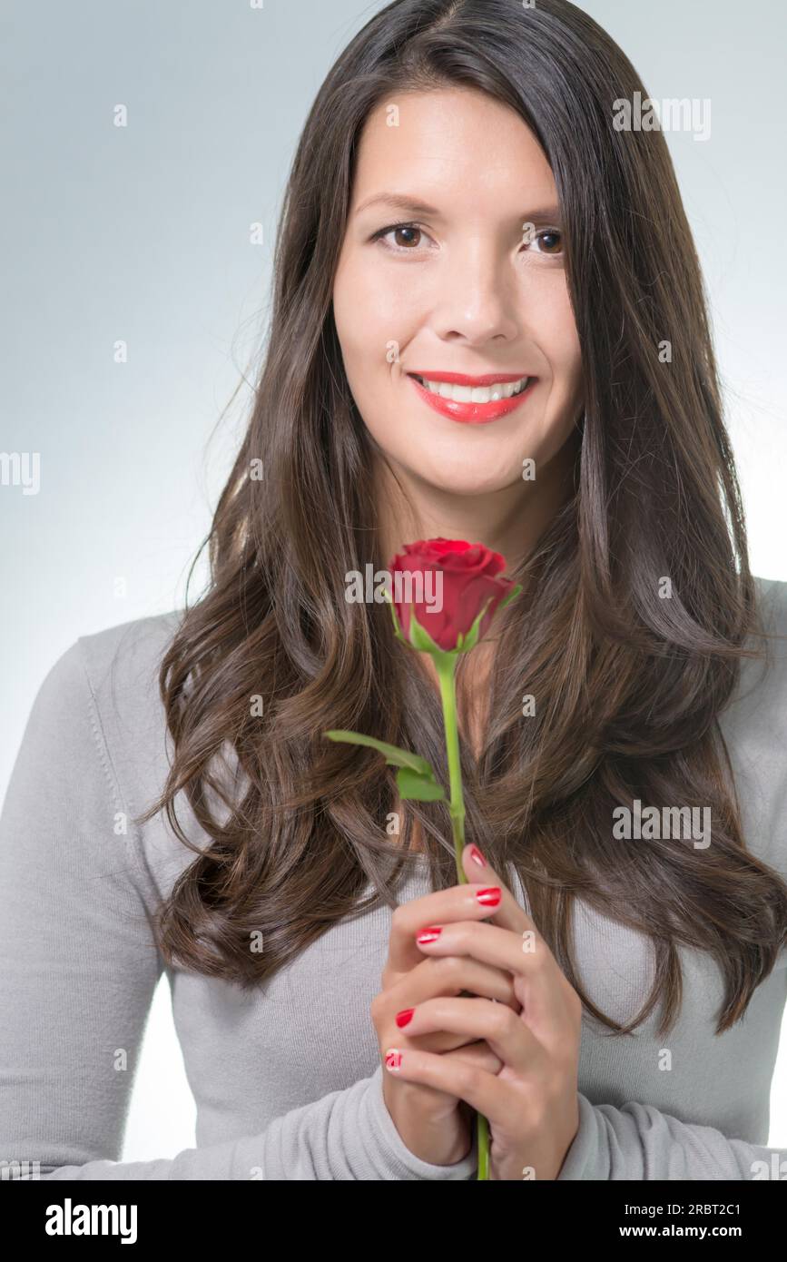 Attraktive Frau mit langem braunem Haar in der Hand einer langstieligen roten Rose, ein Geschenk eines geliebten Menschen zum valentinstag oder zum Geburtstag Stockfoto