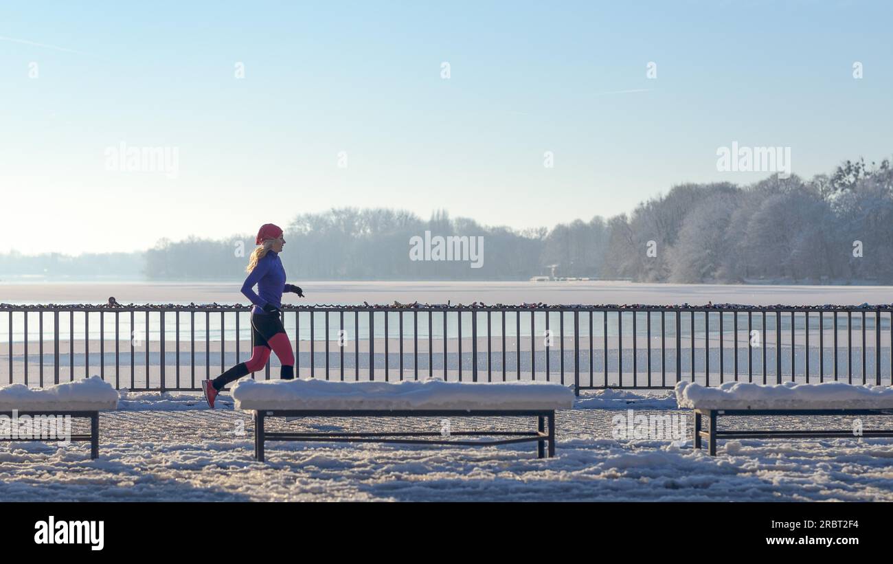 Junge Frau, die im Winterschnee entlang einer Promenade mit Blick auf den Ozean läuft, während sie das belebende kalte Wetter in einem gesunden, aktiven Lebensstil genießt Stockfoto