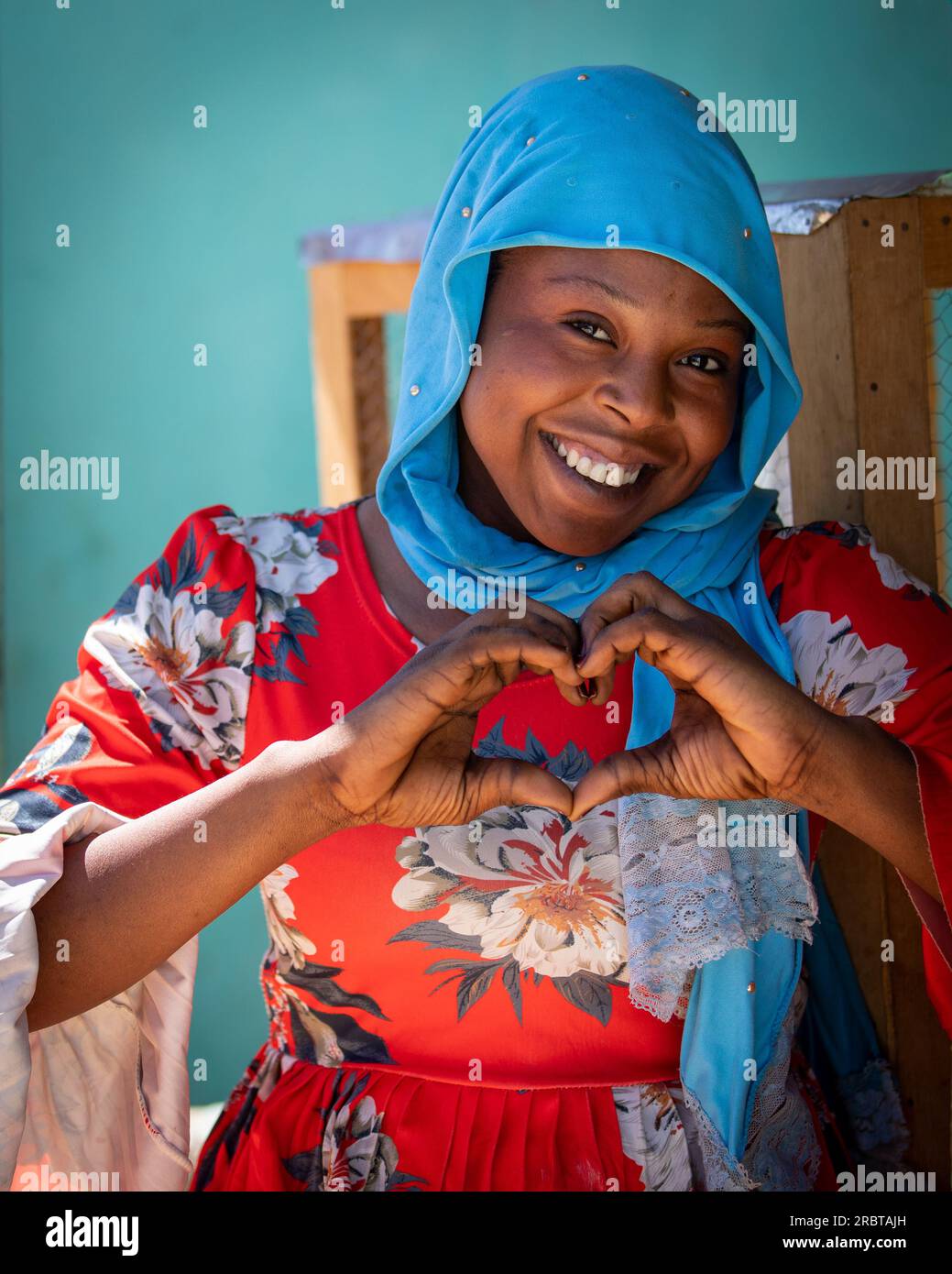 Die charmante tschadische Dame, die in ihrem leuchtend blauen Hidschab strahlt, bildet mit ihren Händen ein herzliches Zeichen Stockfoto