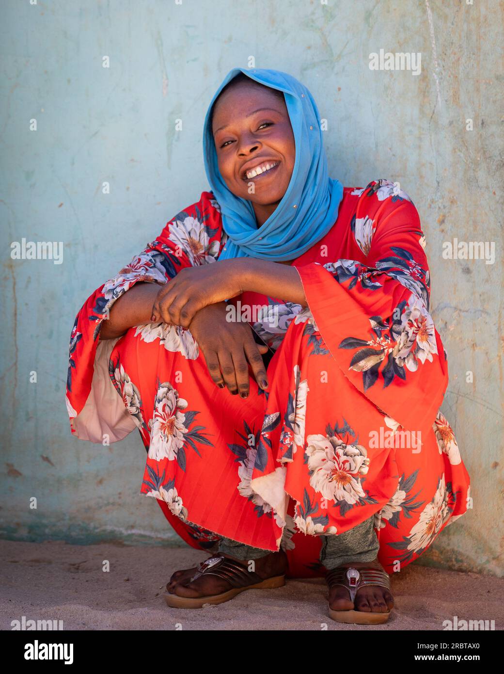 Die fröhliche tschadische Dame in einem leuchtend blauen Hijab schmückt uns mit ihrem strahlenden Lächeln, das an einer Wand sitzt Stockfoto