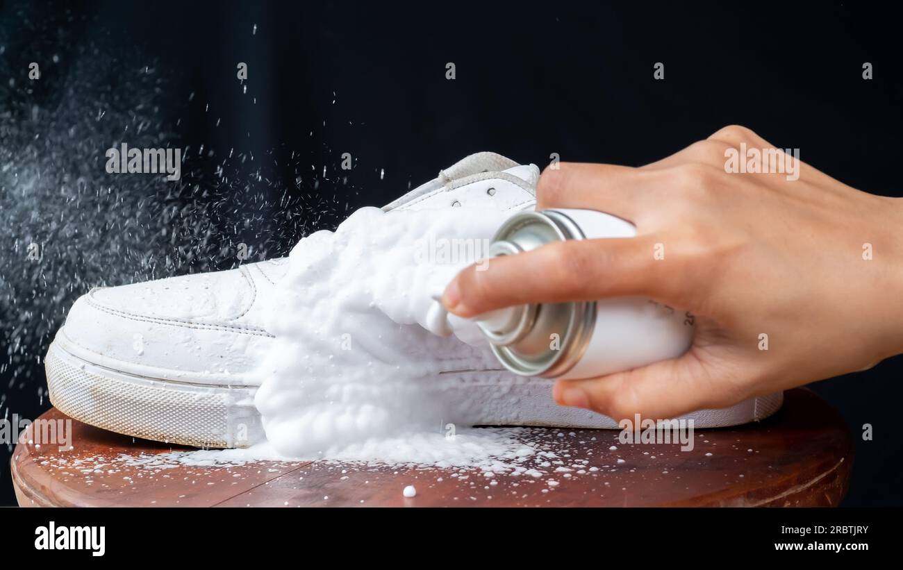 Eine Frau sprüht Reinigungsschaum auf weiße Lederschuhe. Pflege von Lederschuhen. Stockfoto