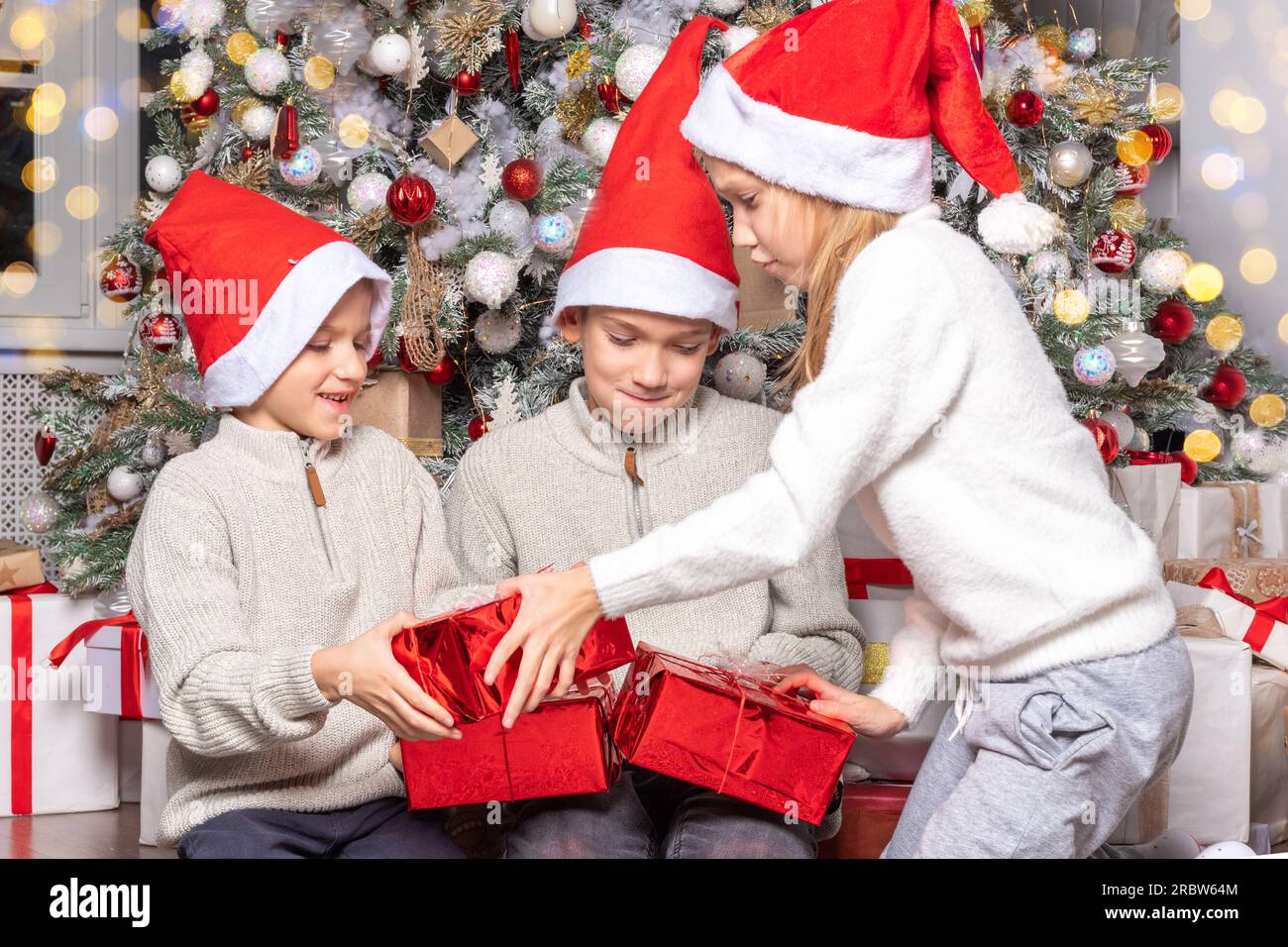 Süße, fröhliche, aufgeregte Kinder, Junge und Mädchen in weihnachtsmannmützen packen weihnachtsgeschenkbox in einem wunderschönen Zimmer aus. Schwester und Bruder schenken Weihnachtsgeschenke in der Nähe von Deko Stockfoto