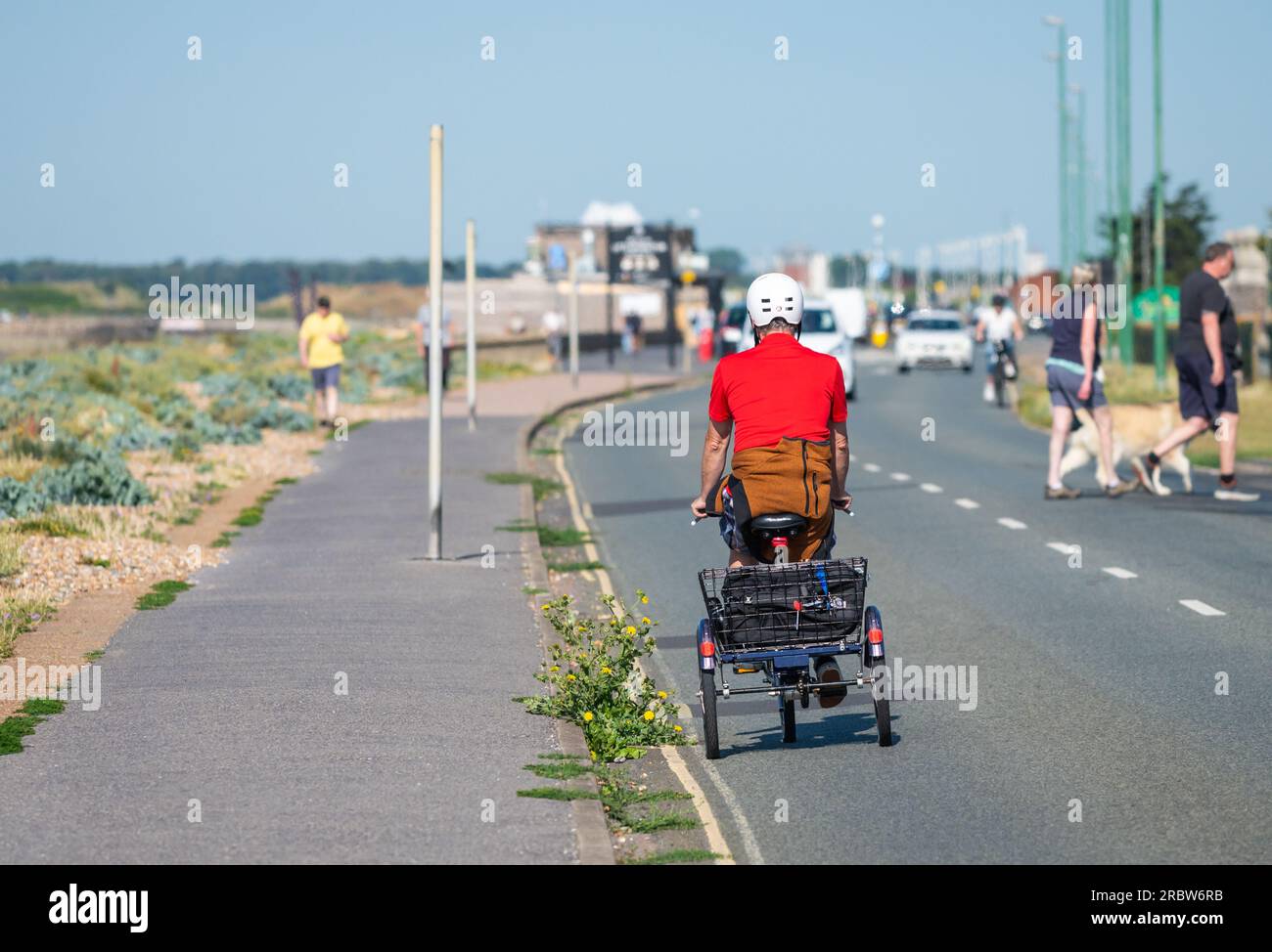 Ein Mann, der im Sommer in Großbritannien mit einem Dreirad, einem 3-rädrigen Motorrad, einem Einkaufskorb und einem Helm auf einer Straße am Meer fährt. Stockfoto