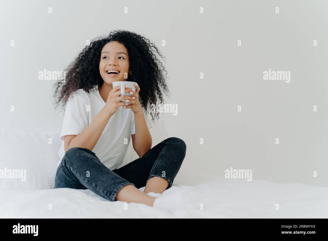 Afro Woman's gemütlicher Morgen: Trinkt ein heißes Getränk im Bett, Blick auf den Inhalt, bequeme Kleidung, sonniges Wetter, weiße Wand. Stockfoto