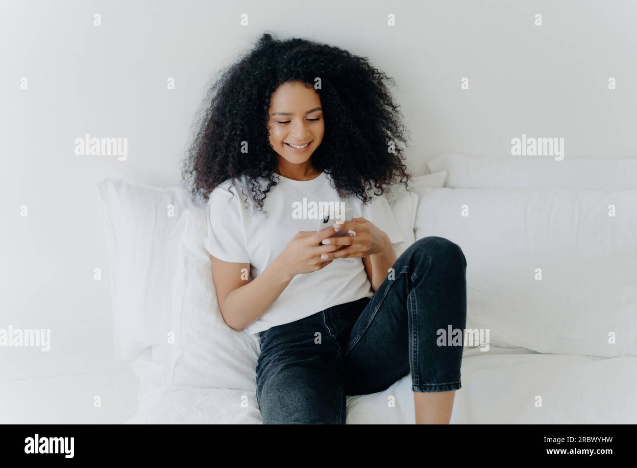 Chill Afro Woman, fröhliches Gesicht, Surfen im Internet am Telefon im gemütlichen weißen Bett, WiFi-Verbindung, häusliche Glückseligkeit. Stockfoto
