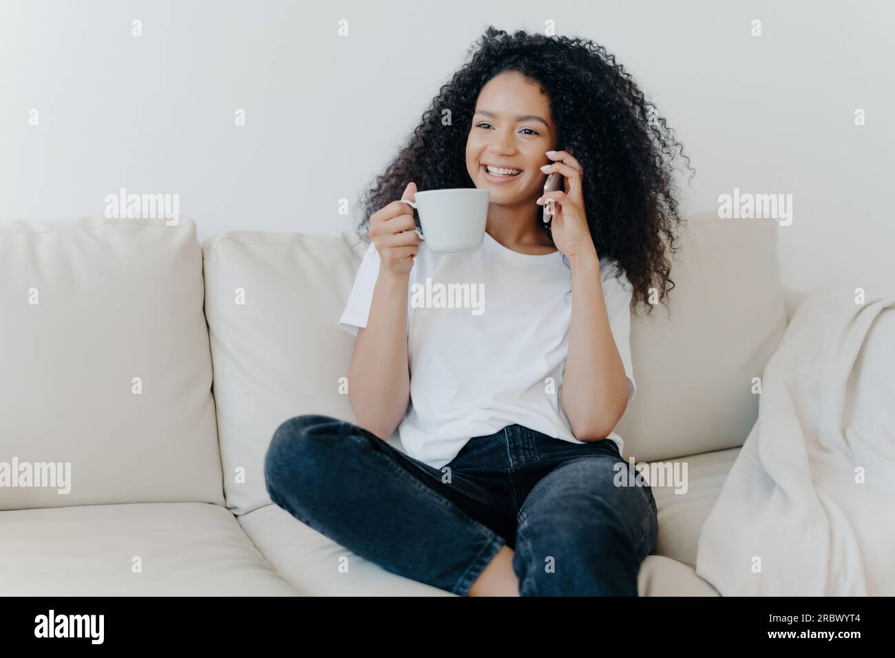 Fröhlicher Afro-Amerikaner auf der Wohnzimmercouch, Kaffeepause, ruft einen Freund mit modernen Geräten an, fröhliches Lächeln, ansprechende Unterhaltung. Tech-Lifestyle-Mischung. Stockfoto