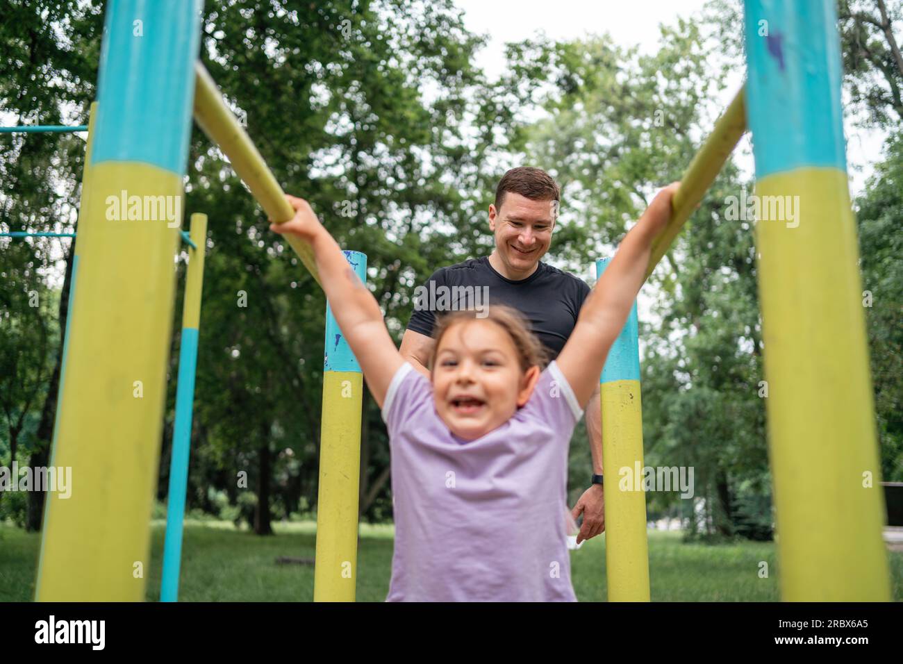 Vater mit Kind, das auf dem Outdoor-Fitnessbereich Pull-ups macht. Gesunder, aktiver Lebensstil, glückliche Familienzeit. Modernes Vaterschaftskonzept Stockfoto