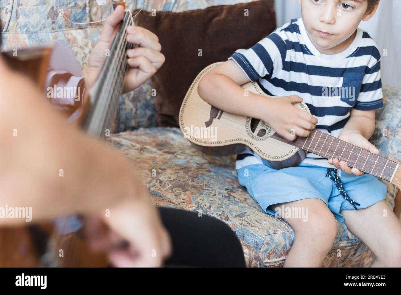 Kaukasisches Kind spielt und macht Musikakkorde mit kleiner Gitarre oder Ukulele, Nahaufnahme. Erwachsene unterrichten das Kind entspannt. Stockfoto