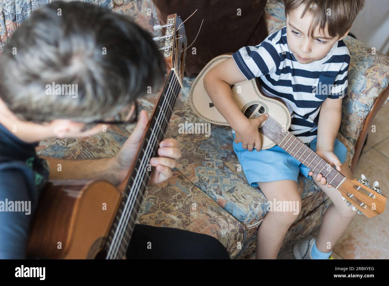 Kaukasisches Kind spielt und macht Musikakkorde mit kleiner Gitarre oder Ukulele, Nahaufnahme. Erwachsene unterrichten das Kind entspannt. Stockfoto