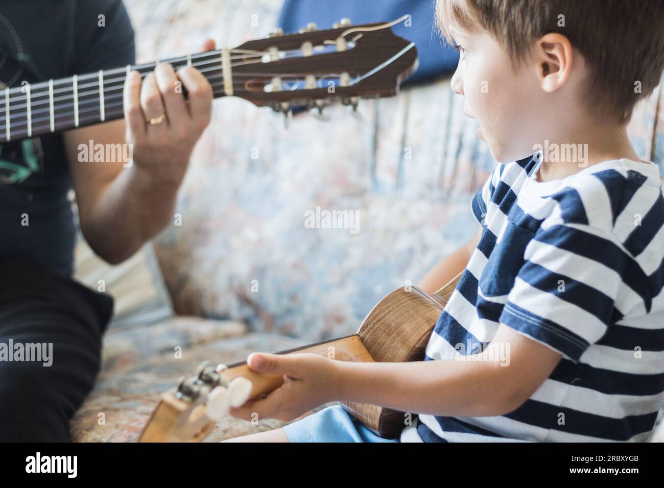 Kaukasisches Kind spielt und macht Musikakkorde mit kleiner Gitarre oder Ukulele, Nahaufnahme. Erwachsene unterrichten das Kind entspannt. Stockfoto