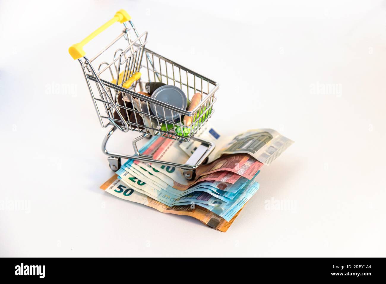 Supermarkt-Einkaufswagen mit wichtigen Waren und mehreren Euro-Banknoten auf weißem Hintergrund. Konzeptionelles Bild der Kaufkraft. Stockfoto