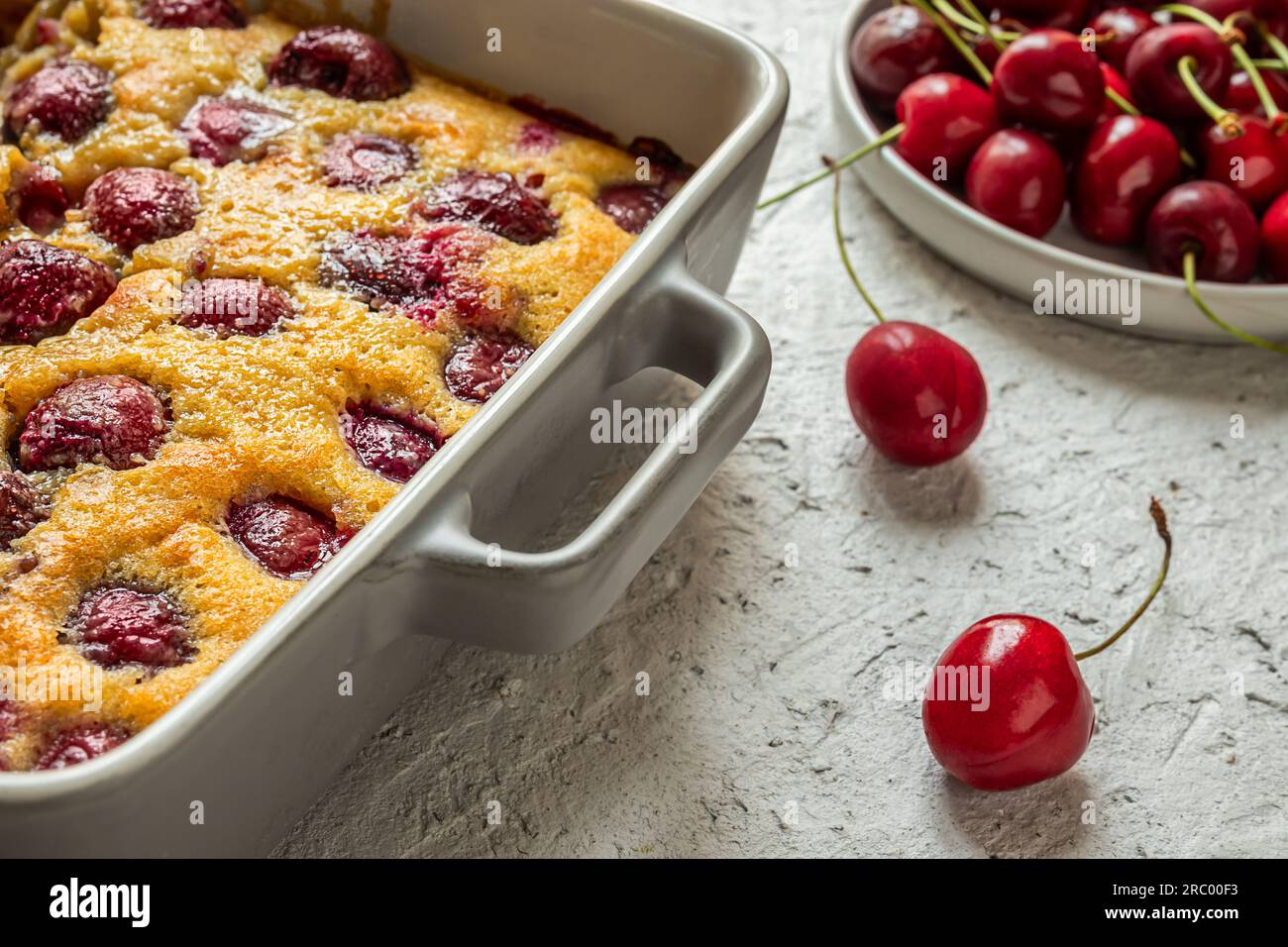 Clafoutis Cherry Torte – traditioneller französischer Kuchen in einer Backform und frische Kirschen auf dem grauen Tisch Stockfoto