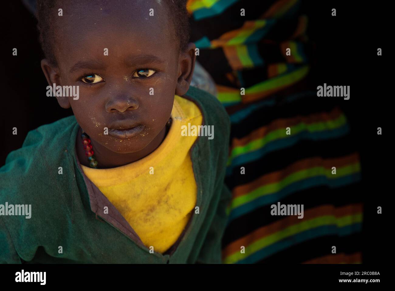 Ein Porträt eines tschadischen Jungen mit Traurigkeit in den Augen, der verwitterte gelbe und dunkelgrüne Kleidung trägt, spiegelt die Widerstandsfähigkeit inmitten sozialer Herausforderungen wider Stockfoto