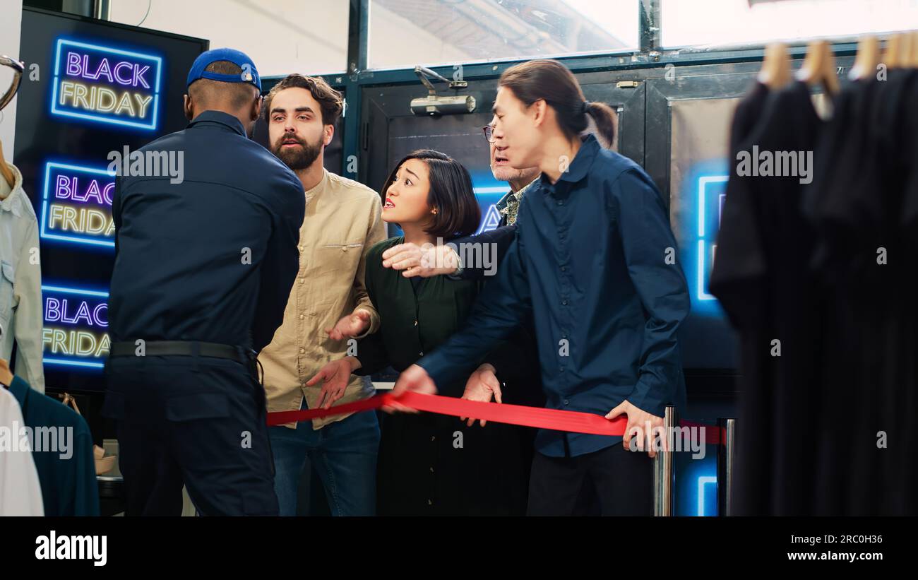 Kunden drängen Bürokratie und betreten einen Bekleidungsladen, um Kleidung auf dem Black friday Sale zu kaufen. Wachmann, der unter Druck mit einer Menge besessener, aggressiver Käufer arbeitet. Handgeführte Aufnahme. Stockfoto