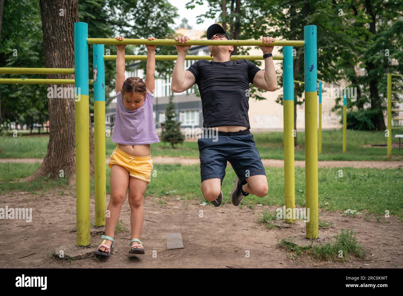 Vater mit Kind, das auf dem Outdoor-Fitnessbereich Pull-ups macht. Gesunder, aktiver Lebensstil, glückliche Familienzeit. Modernes Vaterschaftskonzept Stockfoto
