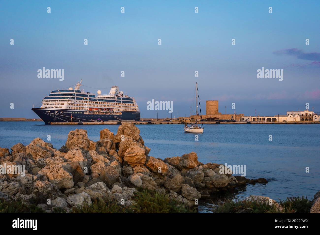 Rhodos, Griechenland - 28. Mai 2023: Sonnenuntergang am Hafen von Mandraki. Ein großes touristisches Kreuzfahrtschiff, das am Pier vor Anker liegt und eine Segelyacht in den Hafen einfährt. Stockfoto