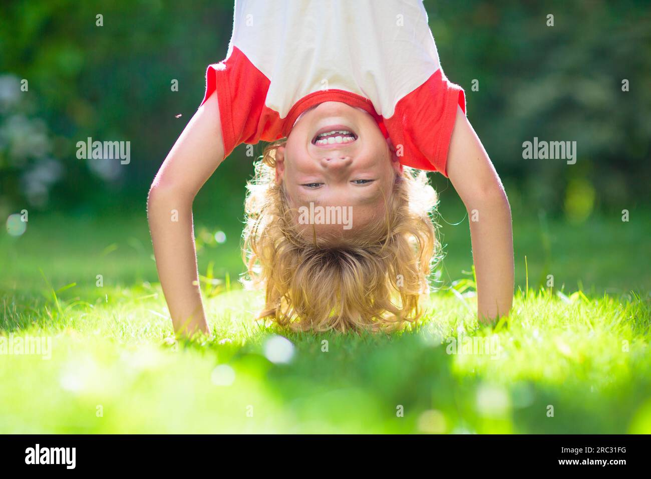 Der kleine Junge hängt kopfüber. Handstand-Übungen für kleine Kinder. Aktives Kind spielt auf sonnigen Gartenrasen. Sommerspaß. Kinder spielen draußen. Stockfoto