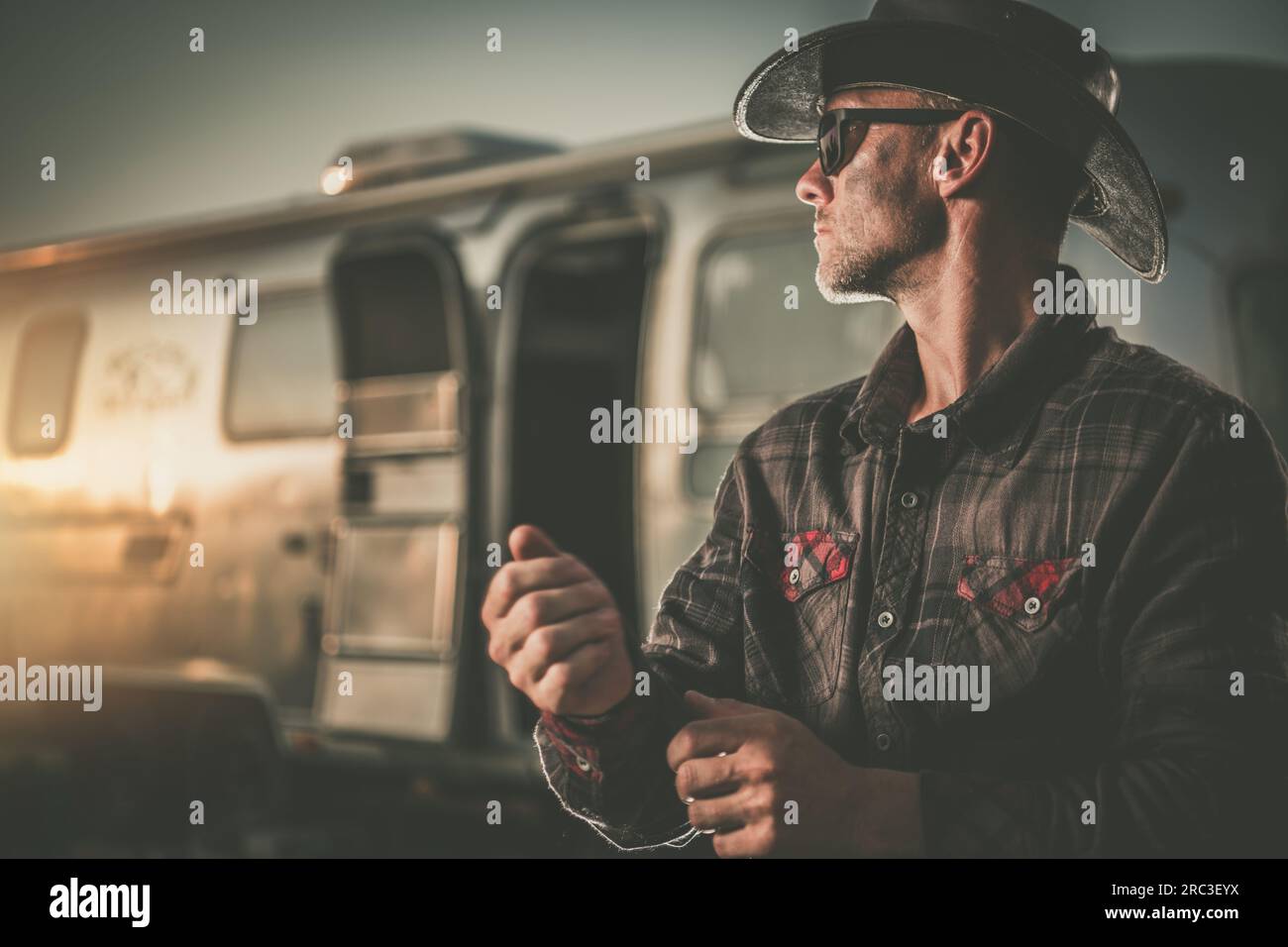 Portrait von American Cowboy in seinem 40s neben seinem Vintage Classic Travel Trailer während Scenic Sunset. Amerikanisches West-Thema Stockfoto