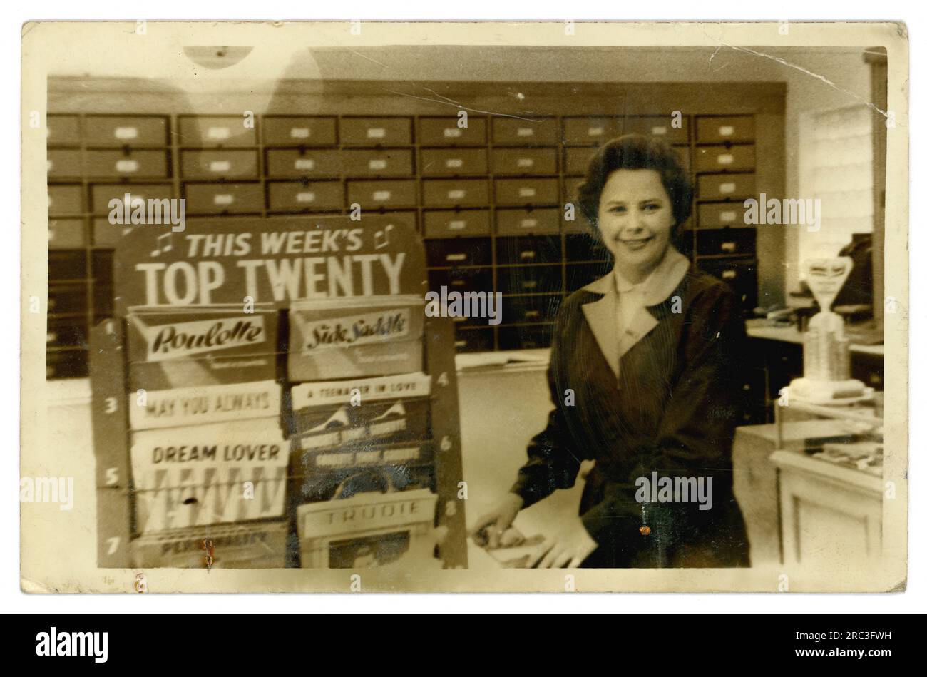Originalfoto aus den 50er Jahren einer attraktiven, lächelnden Ladenassistentin in einem Plattenladen oder Gemischtwarenladen mit einer Plattenverkaufsabteilung, mit dem „Pop Top Twenty“-Ausstellungsstand dieser Woche, Datiert auf 1959 von Ross Conways Roulette, die keine 1 war, und sein Sidesaddle-Hit war auch Nummer eins im Jahr 1959, Großbritannien Stockfoto