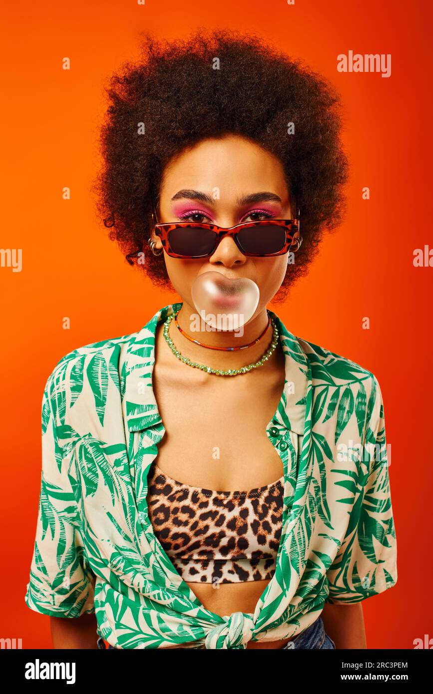 Porträt einer jungen und angesagten afroamerikanischen Frau mit auffälligem Make-up, Sonnenbrille, Sommeranzug und blasendem Kaugummi isoliert auf rot, woma Stockfoto