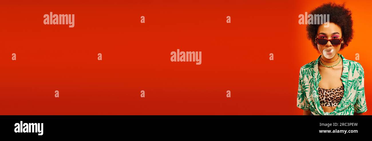 Junge afroamerikanische Frau mit kühnem Make-up, die Sommerkleidung und Sonnenbrille trägt, während sie Kaugummi bläst, während sie isoliert auf Rot steht, Frau mit Stockfoto