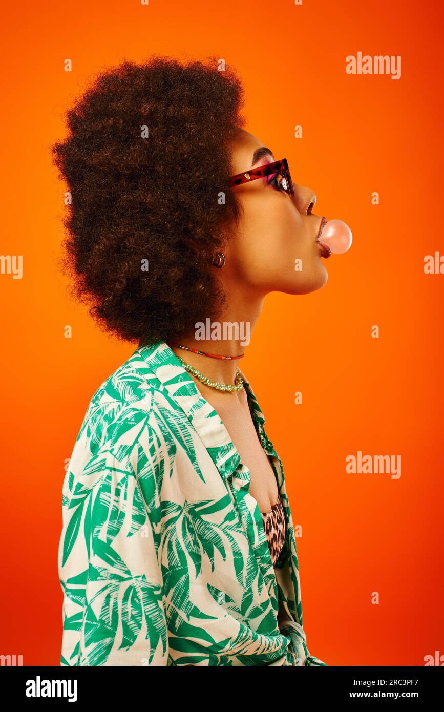 Seitenansicht einer jungen und trendigen afroamerikanischen Frau in Sonnenbrille, die Sommerkleidung trägt, während sie Kaugummi bläst und isoliert auf Rot steht, Frau Stockfoto
