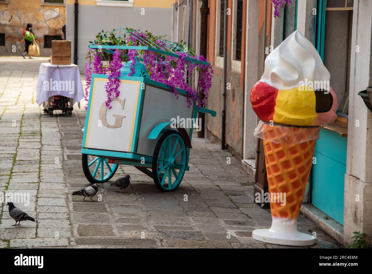 Eine große dekorative Eistüte steht neben einem bunten rollenden Blumenwagen vor einem Geschäft. Stockfoto