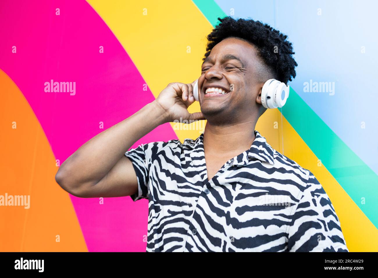 Ein fröhlicher afroamerikanischer Mann in einem Zebrashirt hört Musik mit geschlossenen Augen über kabellose Kopfhörer, während er gegen eine farbenfrohe Wand steht Stockfoto
