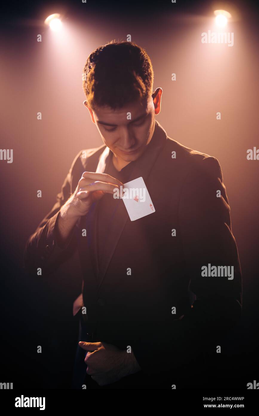 Männlicher Illusionist im Anzug, der einen Zaubertrick zeigt, mit einem Herzchen-Ass, der Karte spielt, während er auf der Bühne gegen die dunkle Wand tritt Stockfoto