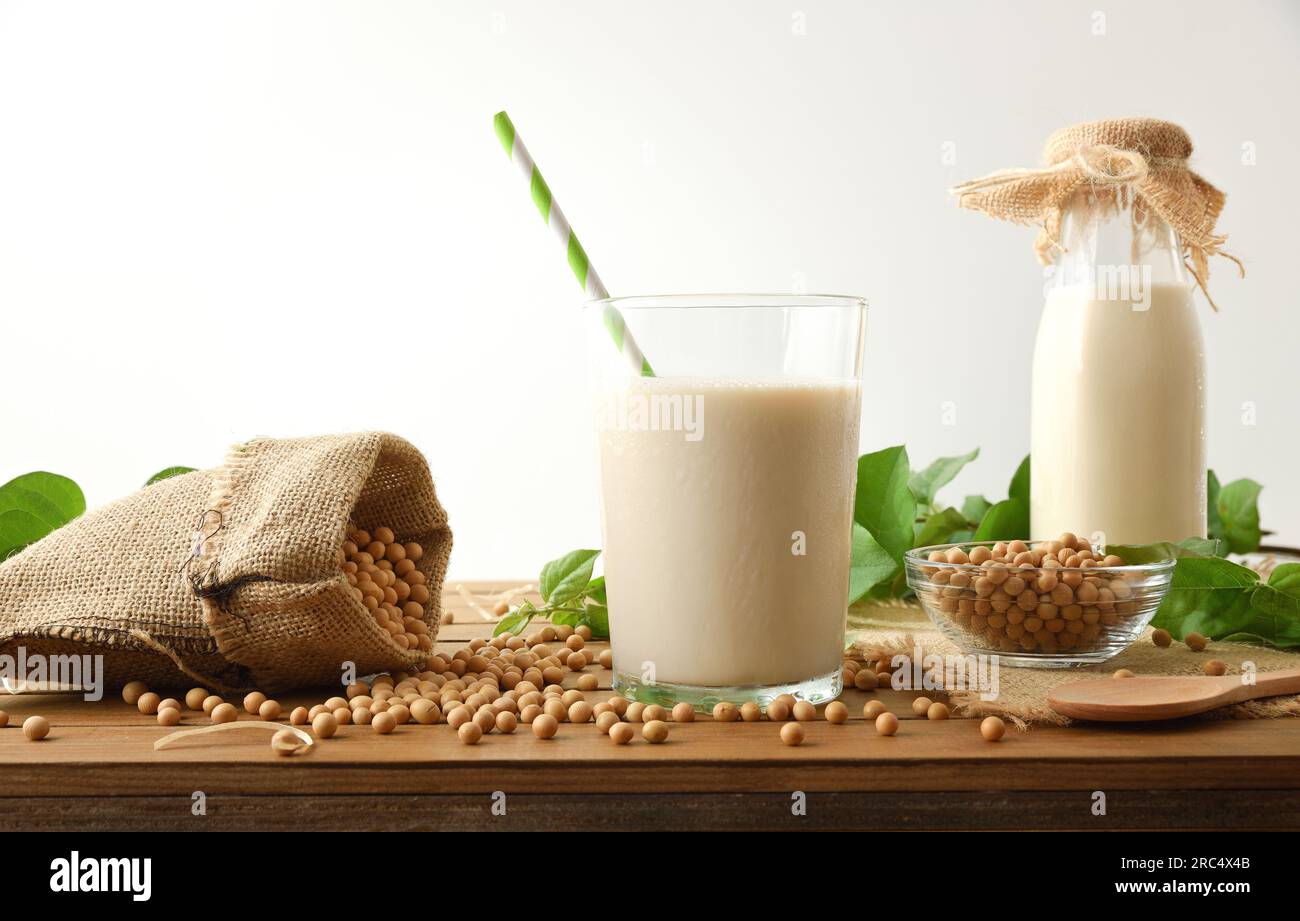 Soja-Getränk in einem Glas mit einem Strohhalm und Körner in einem Stoffsack auf einem Holztisch mit Blättern und einem weißen isolierten Hintergrund. Vorderansicht. Stockfoto