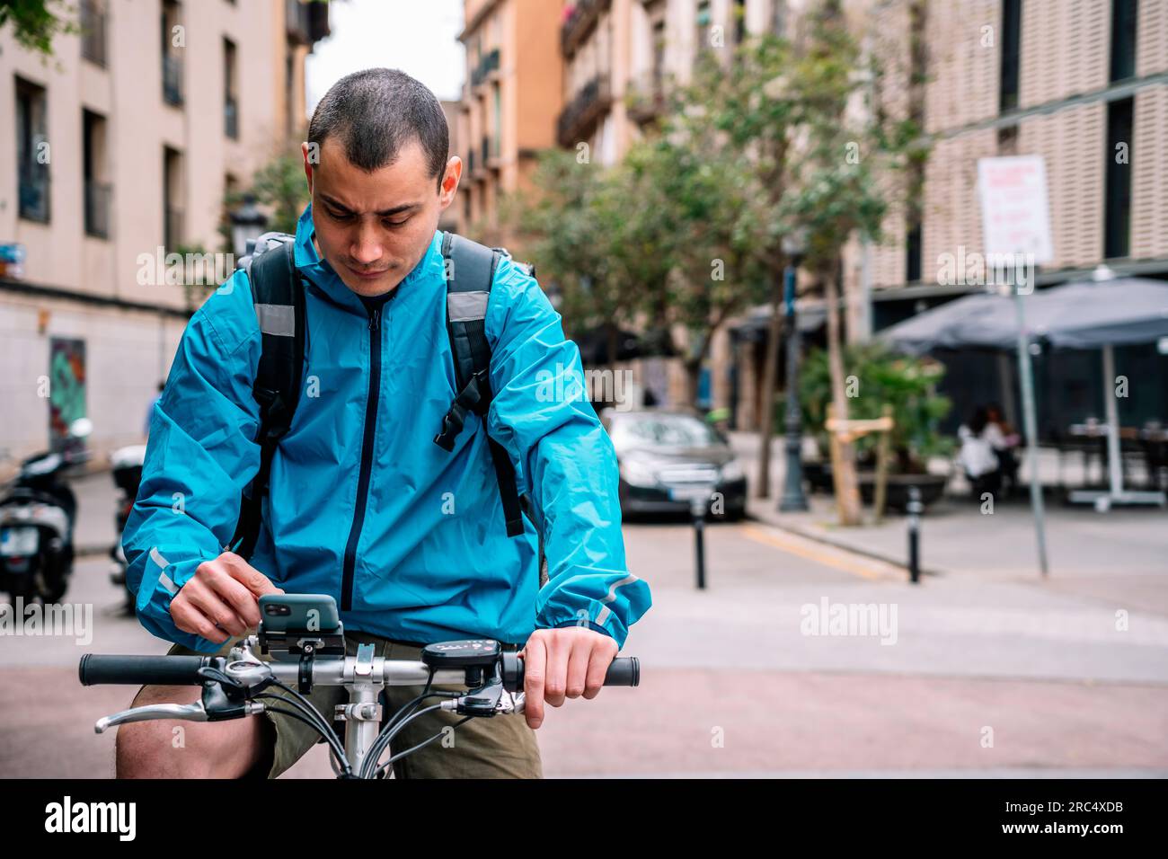 Liefermann in hellblauer Jacke mit Rucksack sitzt auf dem Fahrrad in der Straße der Stadt und telefoniert während der Arbeit in Barcelona Stockfoto
